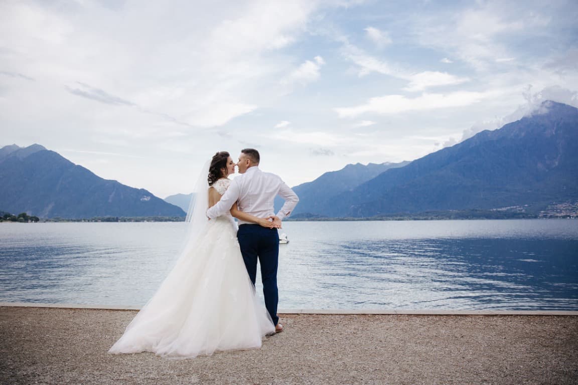 Bride and groom embracing by a lake with mountains in the background